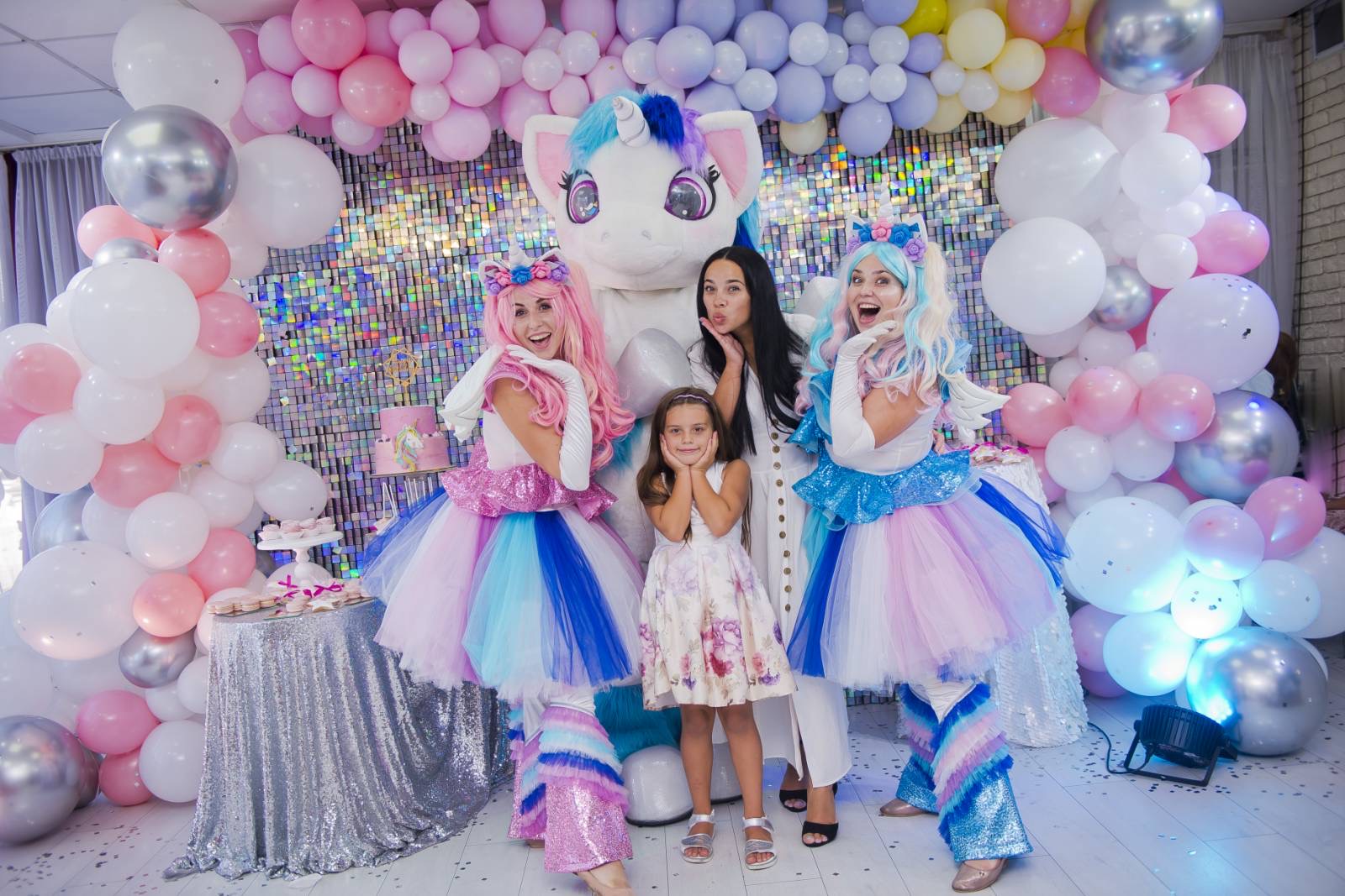 Indoor group photo with unicorn mascot, two pastel-tutu performers, mother, and daughter surrounded by pink and silver balloons.