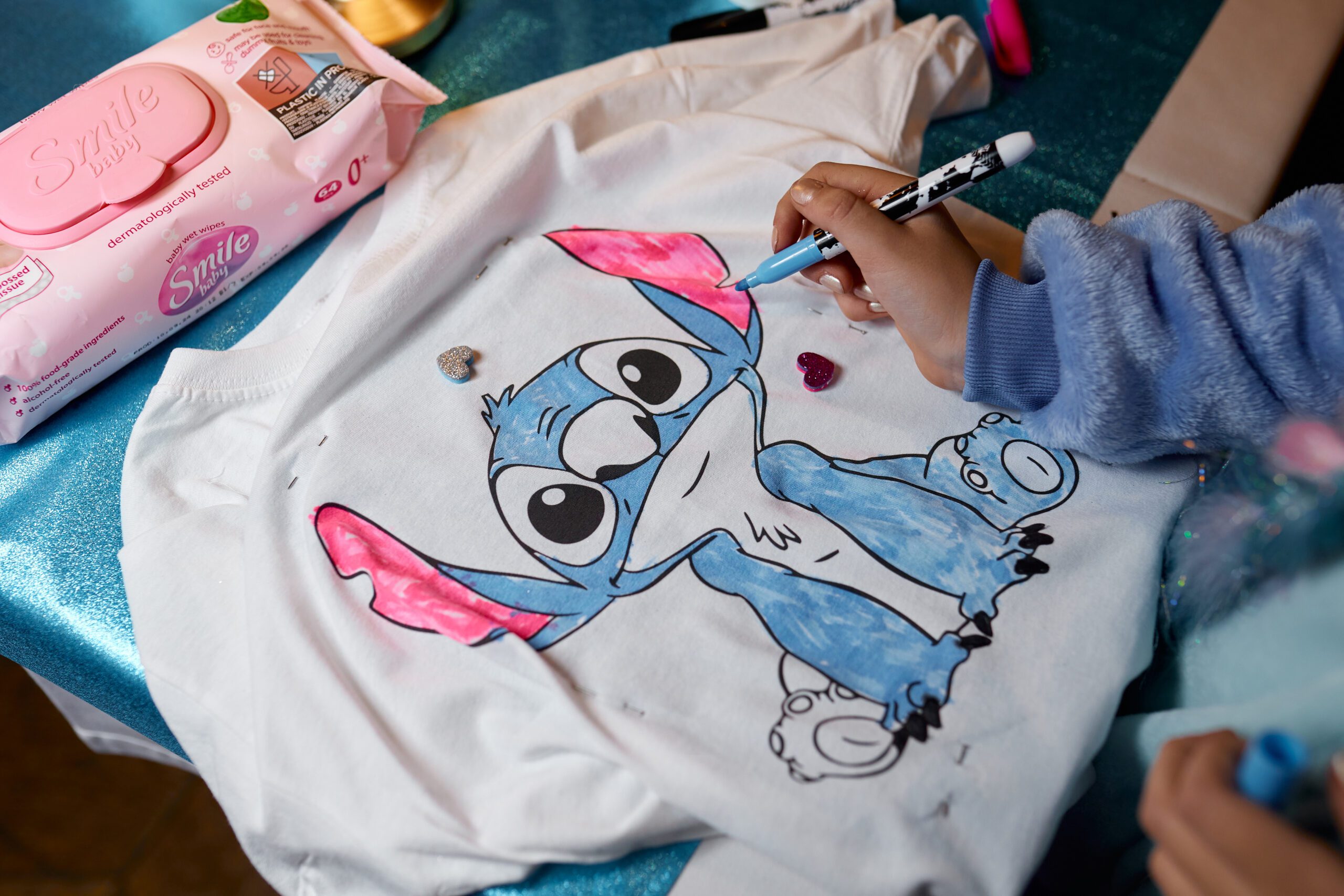 Partially finished T-shirt illustration of a blue cartoon character with pink ears, decorated with glitter heart stickers and fabric markers.