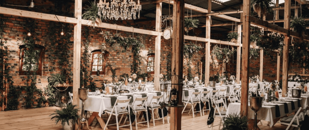Rustic event hall with brick walls, wooden beams, greenery and long banquet tables decorated for a celebration.