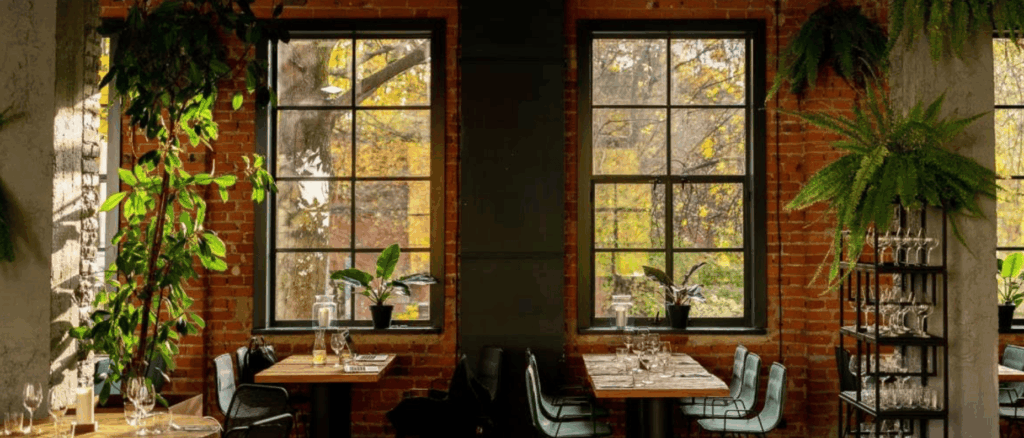 Rustic restaurant with large windows, exposed brick walls, and natural plants creating a warm, urban-jungle vibe.