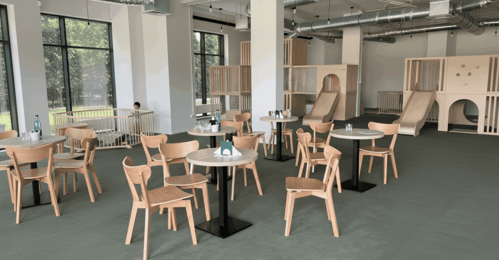 Bright and minimalist family café with natural wooden furniture and a modern indoor playground featuring slides and climbing structures for children.