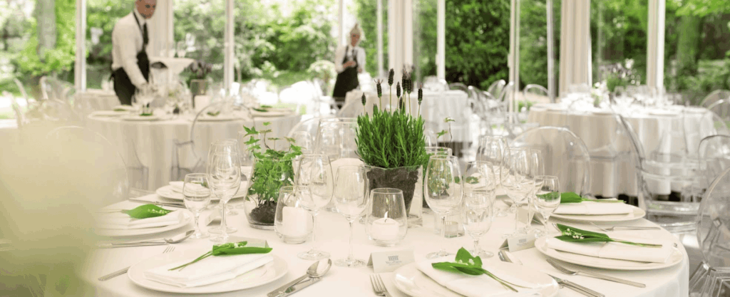 White banquet hall with round tables, clear chairs and fresh green floral accents.