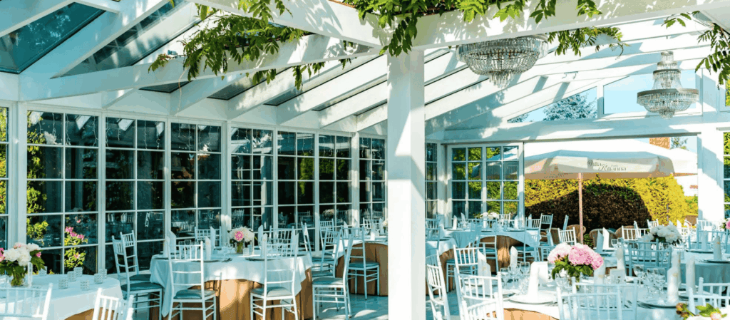 Glass pavilion venue with chandeliers and white table settings for celebrations.